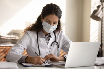 Close up serious professional female doctor wearing protective face mask taking notes, writing in medical journal, filling documents, patient form, sitting at desk with laptop in hospital office