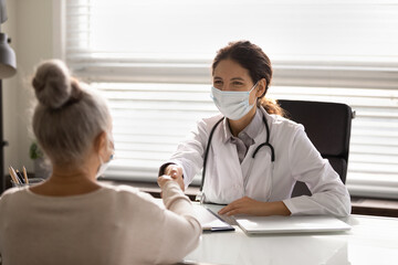 Wall Mural - Close up smiling female doctor wearing face mask and uniform with stethoscope shaking mature patient hand at meeting, elderly woman making health insurance deal, elderly generation healthcare