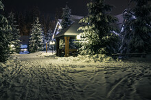 Gazebo, Wooden House And Christmas Trees In The Snow At Night. A Trodden Door Leads To The Gazebo. Lanterns And Garlands Illuminate The Gazebo And The House. Winter Resort In The Mountains