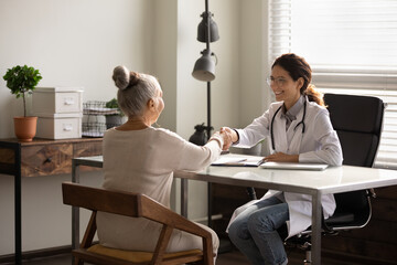 Wall Mural - Smiling female doctor wearing glasses and uniform shaking mature patient hand at meeting in hospital office, celebrating good medical checkup results, senior woman making health insurance deal