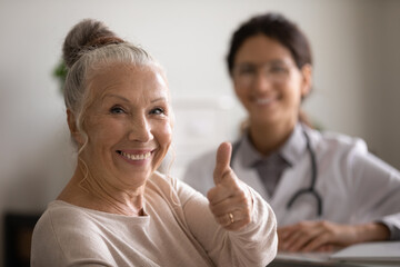 Head shot portrait smiling mature female patient showing thumb up, female therapist and senior man sitting in doctor office, old client satisfied by health insurance or healthcare service concept