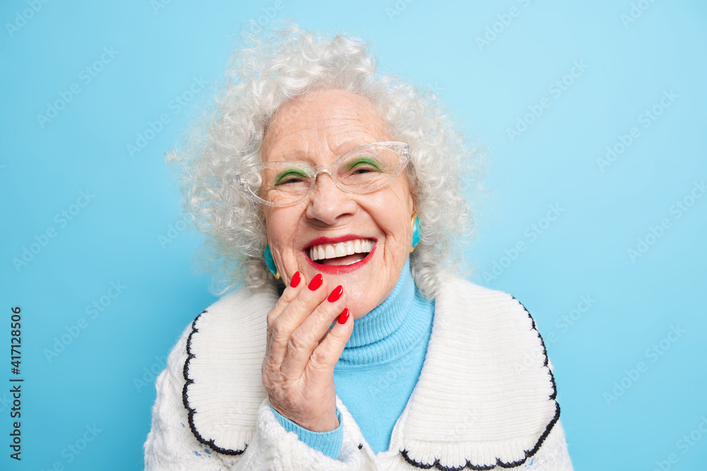 People old age positive emotions concept. Happy grey haired lady smiles broady has white even teeth wears bright makeup dressed in casual sweater happy to spend time with family isolated on blue - obrazy, fototapety, plakaty 
