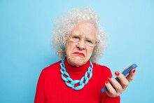 Photo Of Displeased Wrinkled Old Curly Woman Holds Mobile Phone Checks Message Frowns Face Wears Spectacles Red Jumper And Necklace Isolated Over Blue Background. People Age Technology Concept