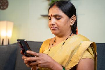 Serious Indian woman busy using mobile phone while sitting on sofa at home - Concept of social media bullying and online messaging.