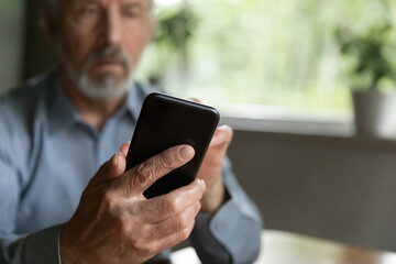 Older 70s aged man using online app on mobile phone, texting message, dialing number for call. Active pensioner looking news on internet or social media. Hand of mature user holding cell. Close up