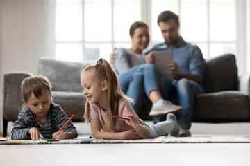 Wall Mural - Happy cute preschooler kids lying on heating floor with carpet in cozy living room, drawing, playing learning games. Family couple watching children, relaxing on sofa, using tablet at home