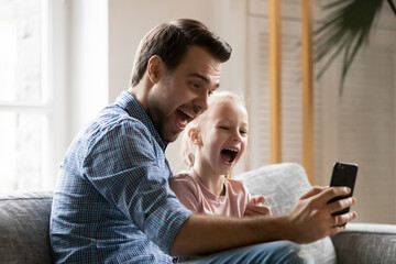 Wall Mural - Excited dad and daughter girl sitting on couch, using mobile phone for video call, shouting and laughing at screen. Happy father and kid having fun together at home, taking selfie on smartphone