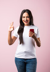 Smiling Indian young woman wears white T Shirt using smartphone or cellphone against pink background