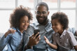 © fizkes - Happy millennial black dad and two cute kids using cell phone for online talk to mom, watching funny video message. Parent and children looking at smartphone screen and laughing on sofa at home.