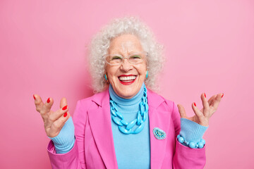 Happy senior curly woman enjoys life in old age raises hands smiles positively dressed in fashionable outfit wears bright makeup has red manicure wrinkled face isolated over pink background.