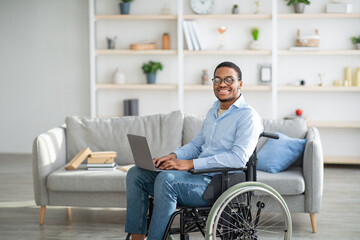 Wall Mural - Happy disabled black man in wheelchair using laptop, working online from home, copy space