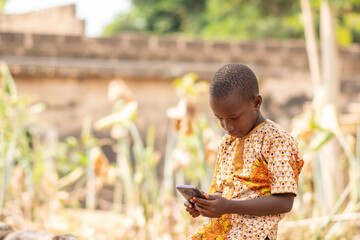 Wall Mural - african kid using a smartphone. african child sitting alone outside viewing something on a phone