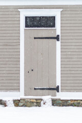 An old wooden and rotten weathered beaten door on the exterior of a tan colour clapboard building with large snowflakes coming down. There are two large wrought iron hinges on the left handed door.