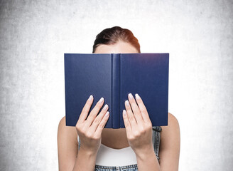 Young woman in casual wear closing her face by blue book. Concrete wall background. Concept of education and distance learning.