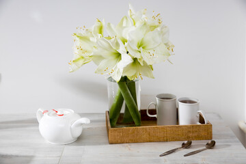 Wall Mural - A bouquet of white lily in a glass vase on a table with two tall cups of coffee, a teapot, spoons.