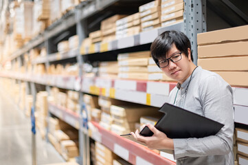 Wall Mural - Young Asian man worker doing stocktaking of product in cardboard box on shelves in warehouse by using clipboard and pen. Physical inventory count concept