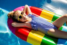 Little Girl In Sunglasses And A Striped Swimsuit Is Lying On An Inflatable Multicolored Striped Mattress In The Pool