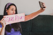 © AliciaFdez - Una mujer morena joven vestida de morado se hace una foto con su teléfono móvil mientras sujeta un cartel con un mensaje feminista 'Girl Power'. Concepto de Día Internacional de la Mujer