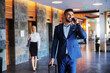 © Dusan Petkovic - Smiling middle-aged man dressed in formal wear walking in the lobby of a fancy hotel and talking on the phone. He is pulling his suitcase.