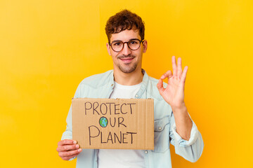 Wall Mural - Young caucasian man holding a protect our planet placard isolated cheerful and confident showing ok gesture.