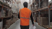 One Man Worker Employee In Orange Vest Walking Along Warehouse With Boxes Of Products With Camera Movement