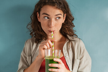 young woman drinking detox juice with straw on blue wall