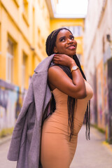 Trendy urban style with a black African girl in a cream colored dress on a city street. Posed with a jacket on his shoulder