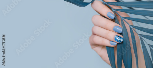 Female hand with blue nail design. Blue nail polish manicure and glitter golden nail art. Female hand hold tropic leaf