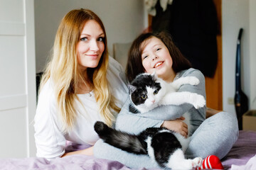 Lovely young mother and cute school kid girl cuddling together with cat in bed in morning. Happy family of young single woman and beautiful daughter, indoors, getting ready and morning routine.