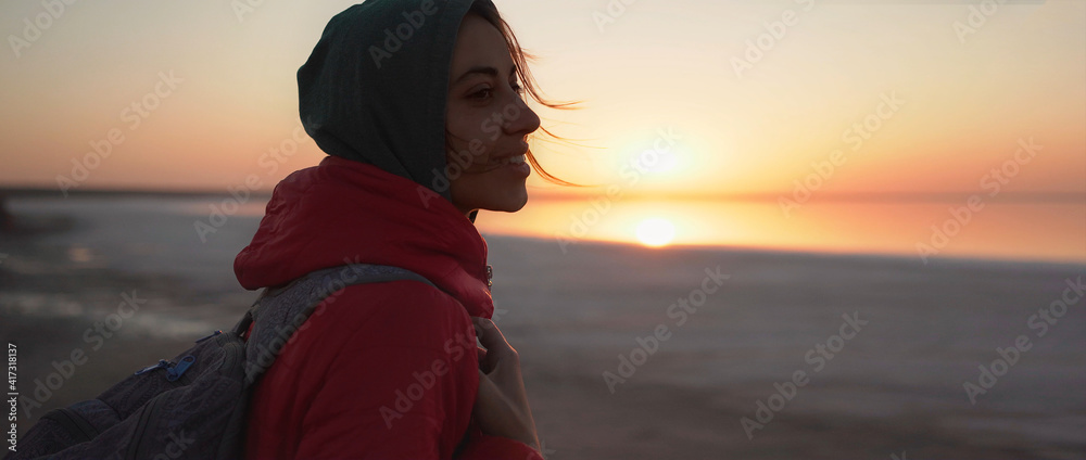close up portrait woman traveler looking at sunrise at sea beach. girl in red jacket and hoodie with wind blowing golden hair from sunlight is enjoying scenery landscape - obrazy, fototapety, plakaty 