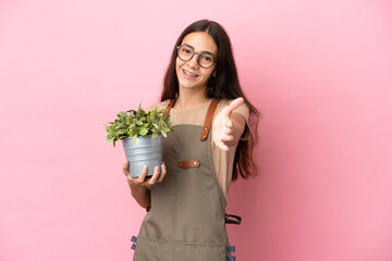 Young gardener girl holding a plant isolated on pink background shaking hands for closing a good deal