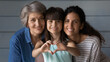 © fizkes - Close up family portrait of happy three generations of Hispanic women pose together show love heart hand gesture. Smiling little Latino girl child with young mother and senior grandmother feel united.
