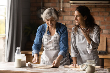 Overjoyed adult Hispanic girl with senior mom make dough bake cookies pastries together on weekend. Happy millennial Latino woman with mature mother have fun cooking dessert at home kitchen.