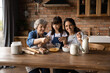 Happy three generations of Hispanic women gather in kitchen cook delicious breakfast together. Smiling little girl with young mom and mature grandmother prepare pancakes or bake at home on weekend.
