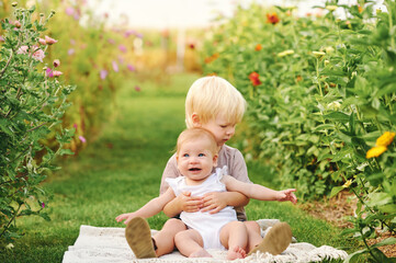 Wall Mural - Outdoor portrait of adorable toddler boy holding his baby sister, kids in beautiful garden