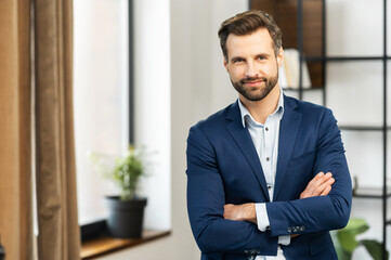 Successful confident young bearded brunet adult male entrepreneur businessman professional in business casual clothes standing with arms crossed folded at the modern home office and looking at camera