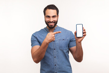 Wall Mural - Happy cheerful beaded man in blue shirt pointing finger at smartphone with empty screen looking at camera with toothy smile, freespace for adv. Indoor studio shot isolated on white background