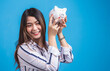 © Pituk - Smiling happy young woman holding a piggy bank in blue background.
