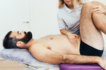 Wall Mural - Bearded male patient lying on the stretcher in the physiotherapy clinic is treated for abdominal pain by the female alternative medicine specialist.