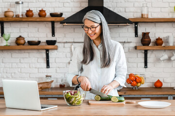 Cheerful caucasian middle-aged woman cooking in the kitchen using laptop