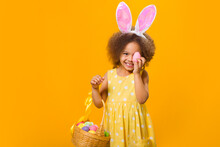 A Cheerful African Girl With Rabbit Ears On Her Head With A Basket Of Colored Eggs In Her Hands.