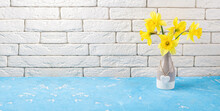 Spring Flowers Yellow Daffodils Bouquet In Vase On Blue Table, White Brick Wall. Yellow Narcissus Or Daffodil Flowers. Spring Holidays, Mothers Day, Easter Concept. Copy Space
