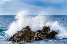 Wave Crashing Against Rock Near The Shore In Laguna Beach, California. White Spray In The Air; Blue Pacific Ocean, Flying Cormorant In The Background. Blue Sky And Clouds Above.
