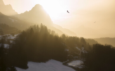 Wall Mural - epic glowing sunset in the mountains with birds circling over church village - sahara dust