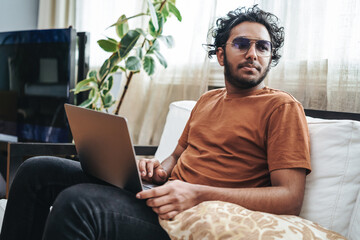 leisure activity of a indian guy with sunglasses dressed in casual attire posing on sofa with a lapt