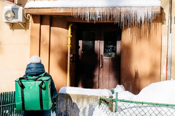 Rear view of caucasian delivery woman wearing warm clothes with green delivery backpack delivers on foot takeaway food in winter snowy cold day city street.