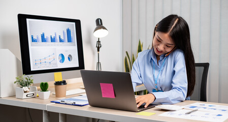 Asian businesswoman talking on the phone and working on a laptop with a cheerful and happy smile while working at the office