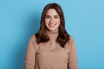 Close up portrait of beautiful young European woman model with dark hair looking at camera with charming cute smile while posing against blue wall, attractive happy female expressing positive.