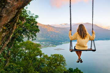 Wall Mural - Summer vacation. Young woman sit on tree rope swing on high cliff above tropical lake. Happy girl looking at amazing jungle view. Buyan lake is popular travel destinations in Bali island, Indonesia