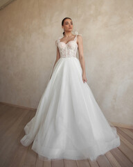 Full length portrait of young beautiful woman wearing white wedding dress. Elegant bride standing and posing against beige wall